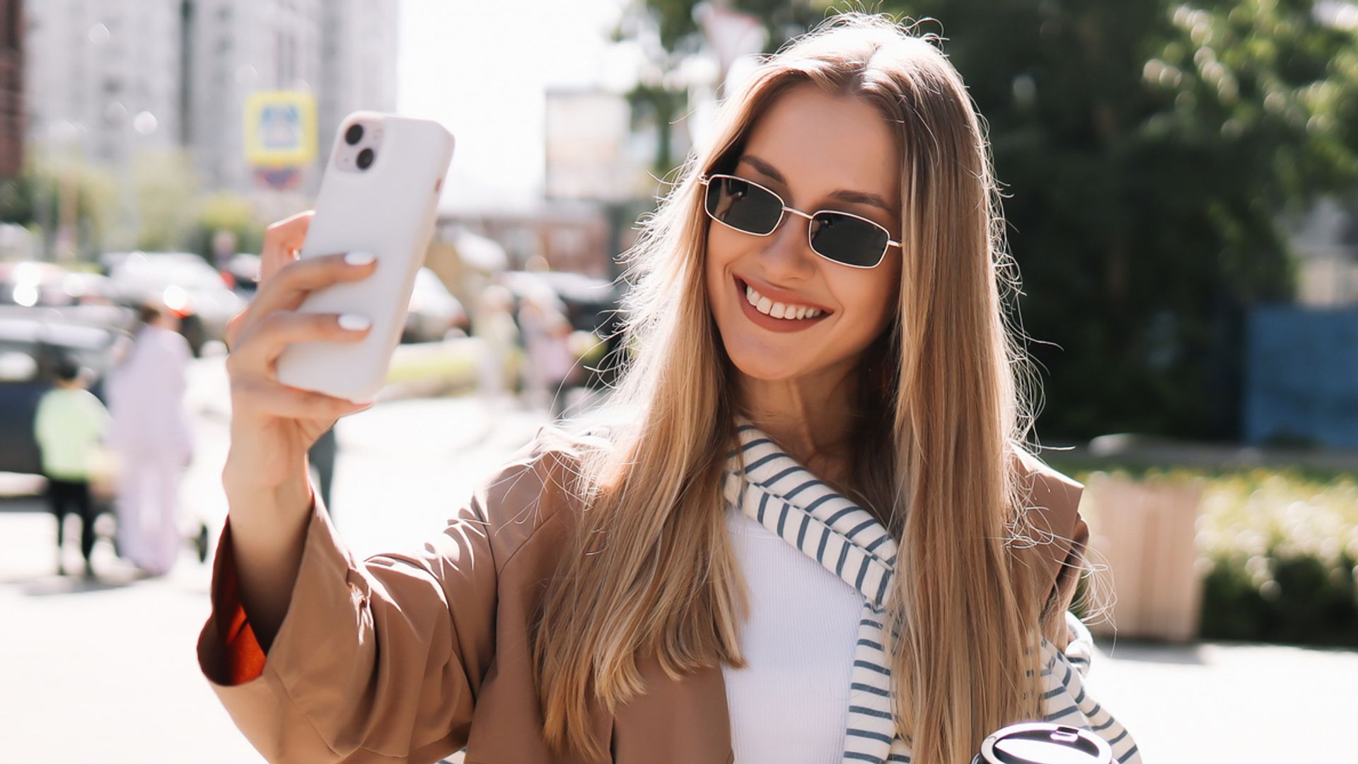 Portrait of a smiling young blonde woman in sunglasses and fashionable clothes walking through the streets of the city holding a glass drinking hot coffee outdoors. A beautiful millennial student girl communicates using a mobile phone and wireless technology takes selfies and laughs while looking at the camera