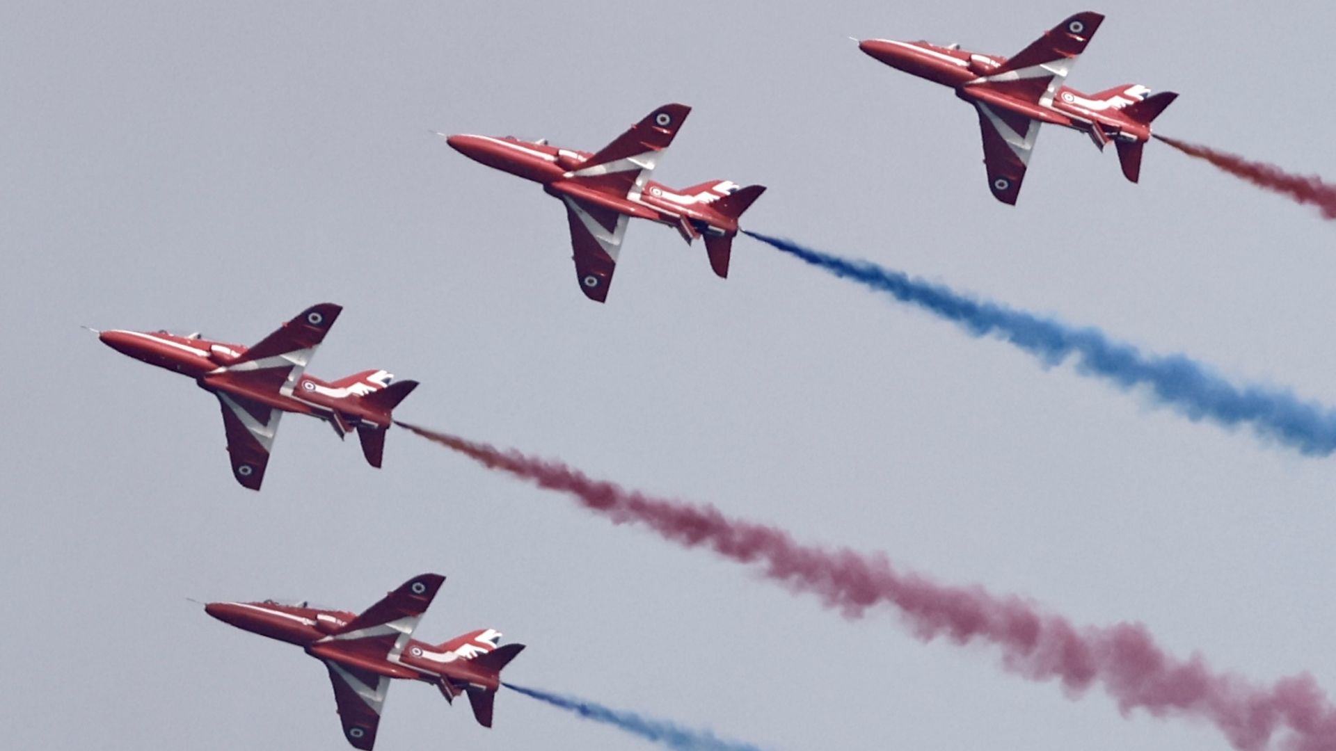 Four red arrows shoot across the sky with blue and red smoke coming from their tails