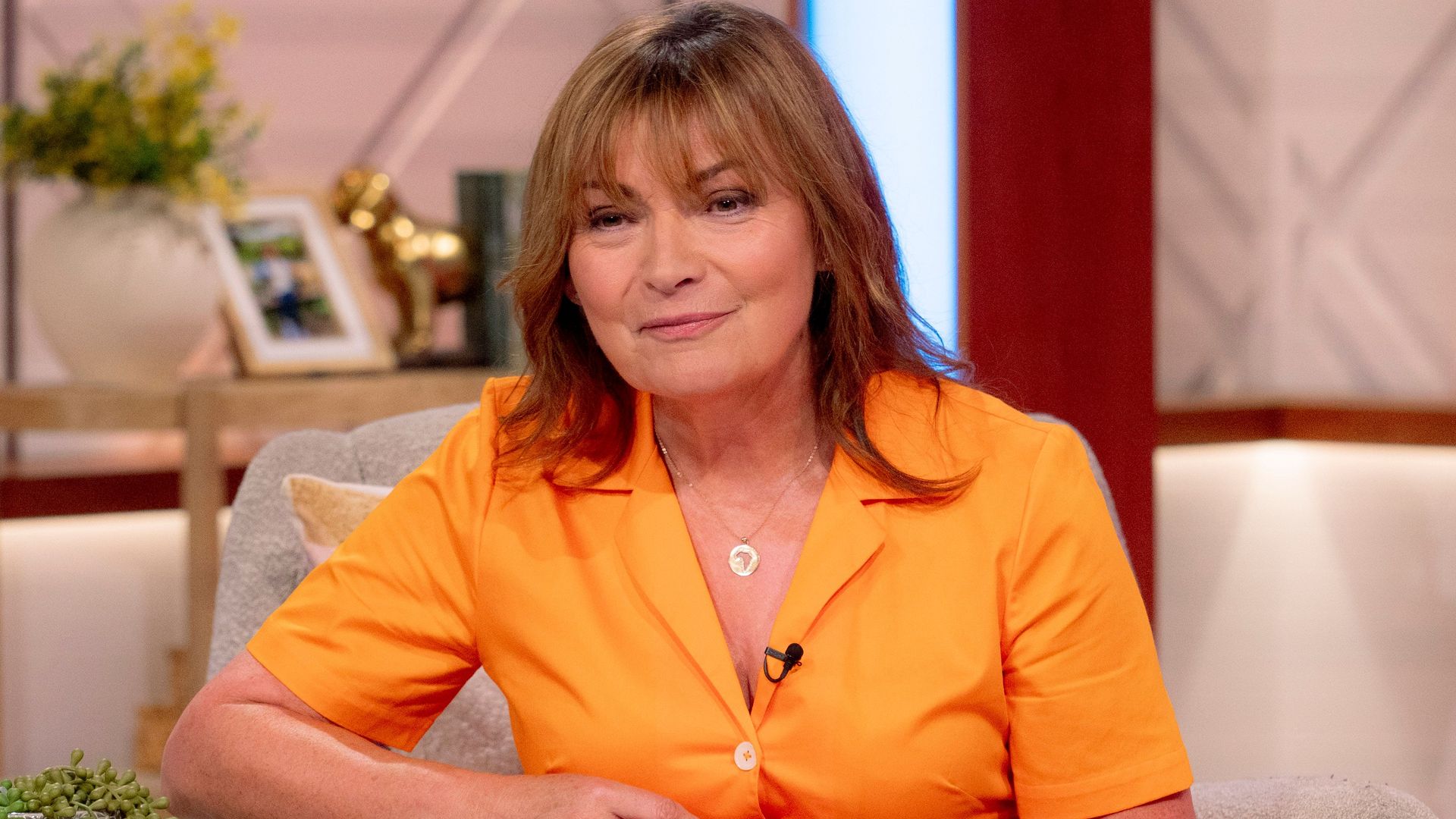 Lorraine Kelly sits on sofa wearing orange shirt