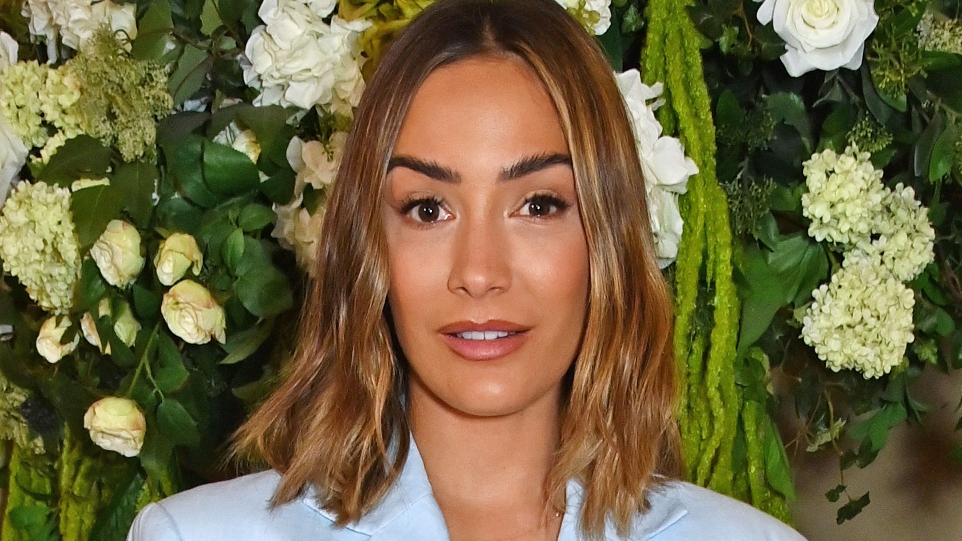 Frankie Bridge in a blue suit standing in front of white flowers