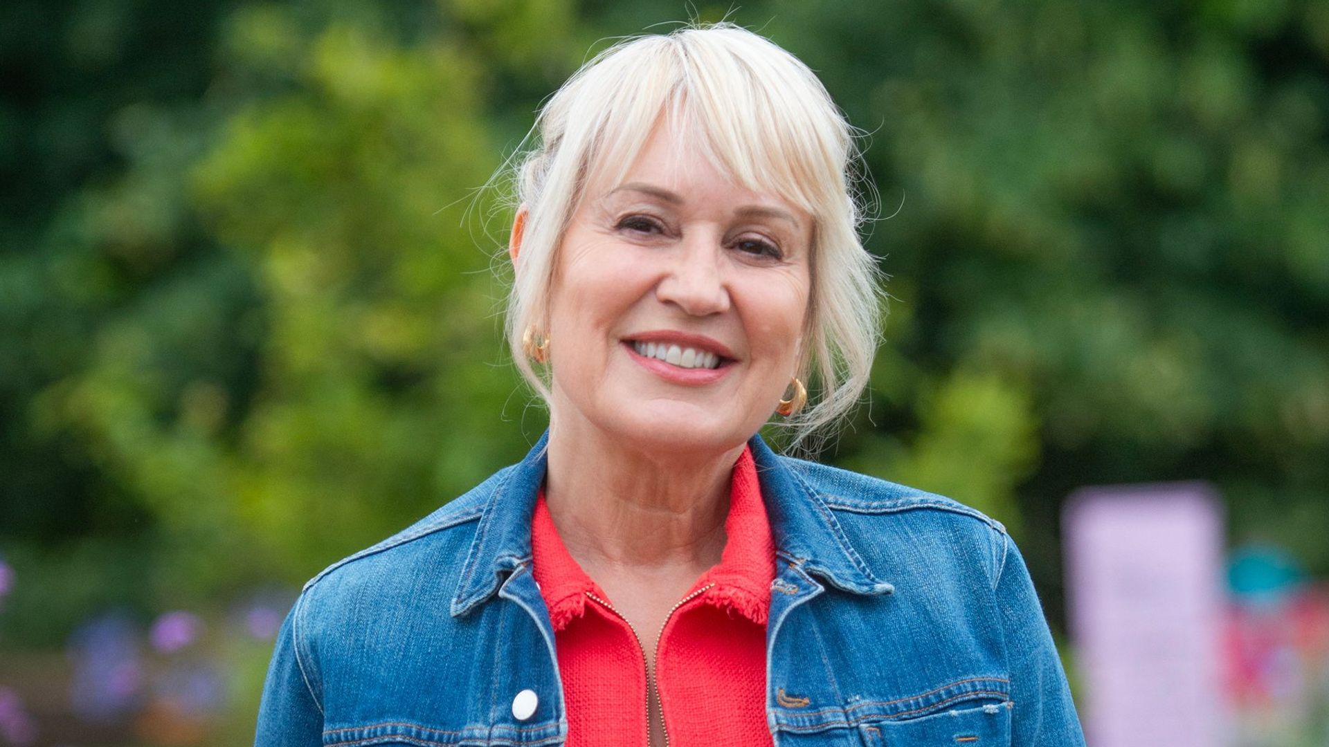 Nicki Chapman in red jumpsuit and denim jacket smiling for camera outside while hosting garden festival coverage