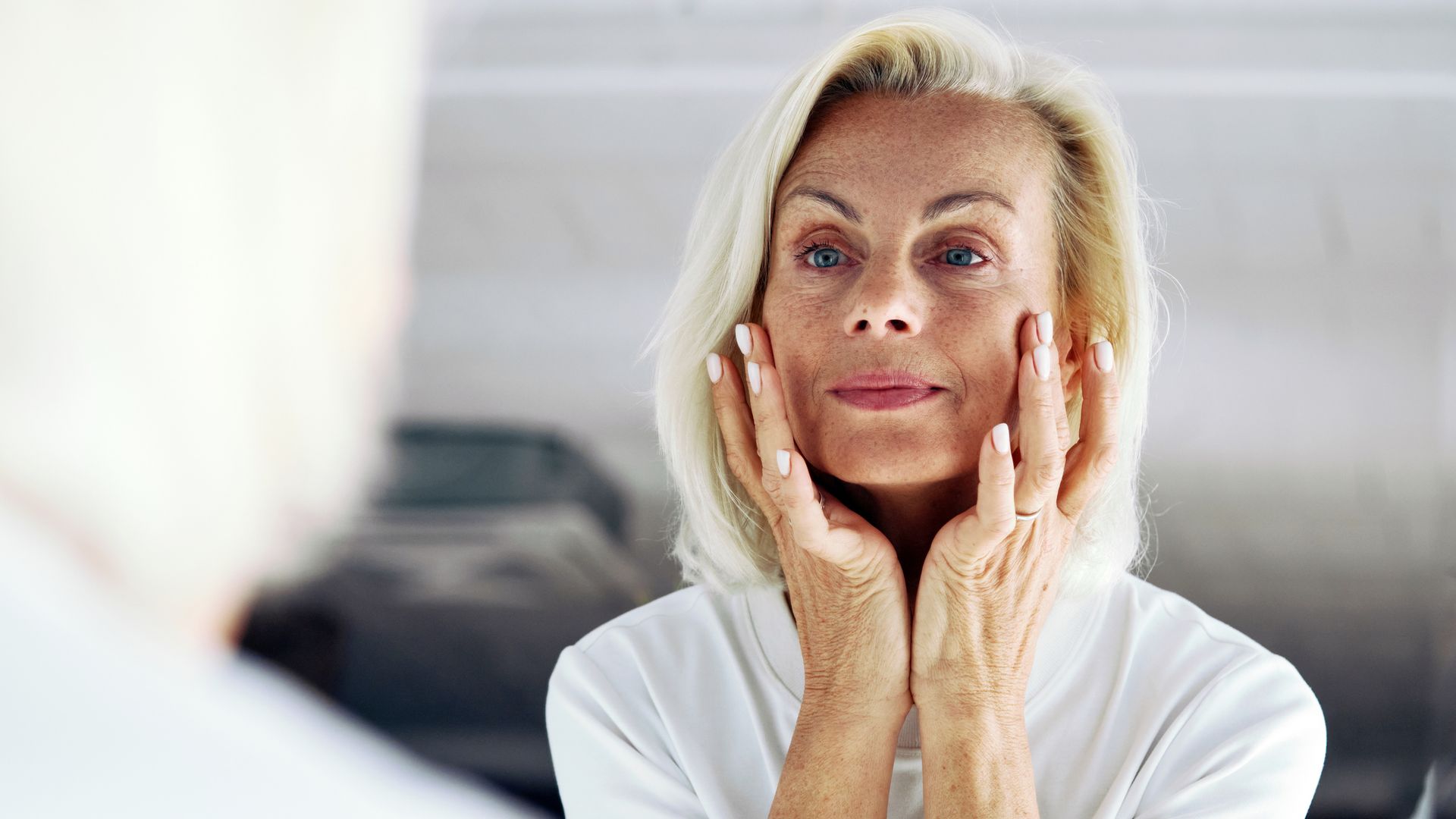 Older woman standing in front of mirror, gently touching her face with hands, close up