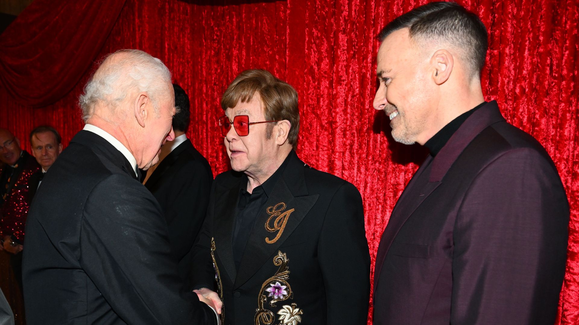 King Charles shaking hands with Elton John as David Furnish looks on