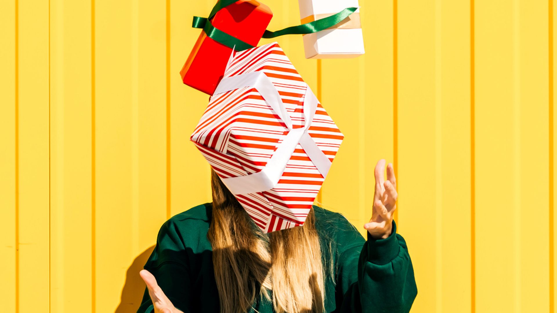 Woman in Christmas hat and green sweater catching Christmas gift boxes against the yellow wall. Funny summer Christmas and sales concept.