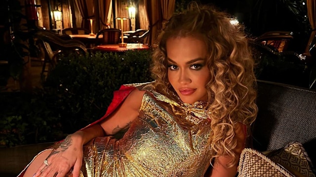 Rita Ora lounges elegantly in a luxurious, textured gold dress