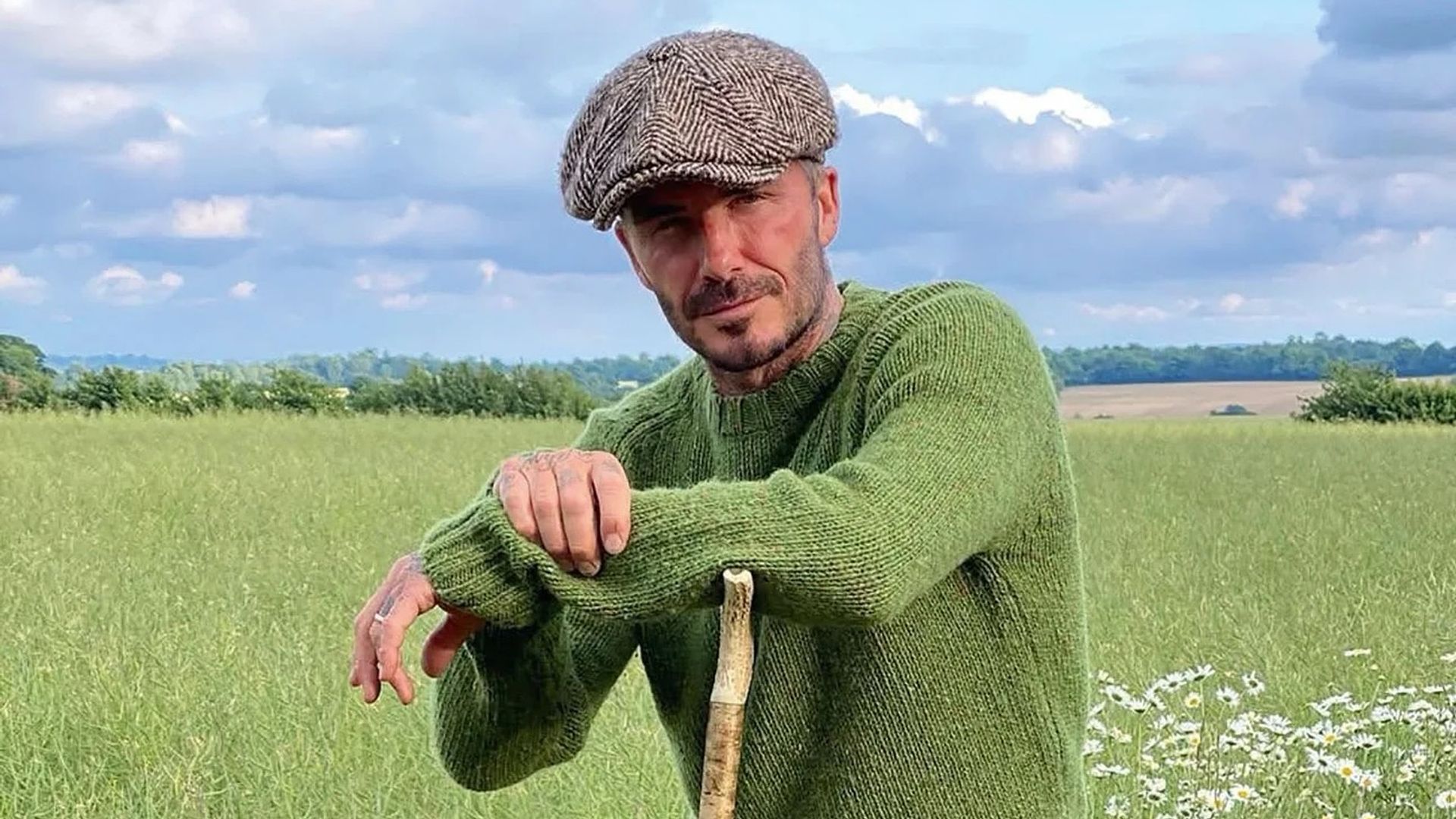 David Beckham wearing green jumper and flat cap standing in field