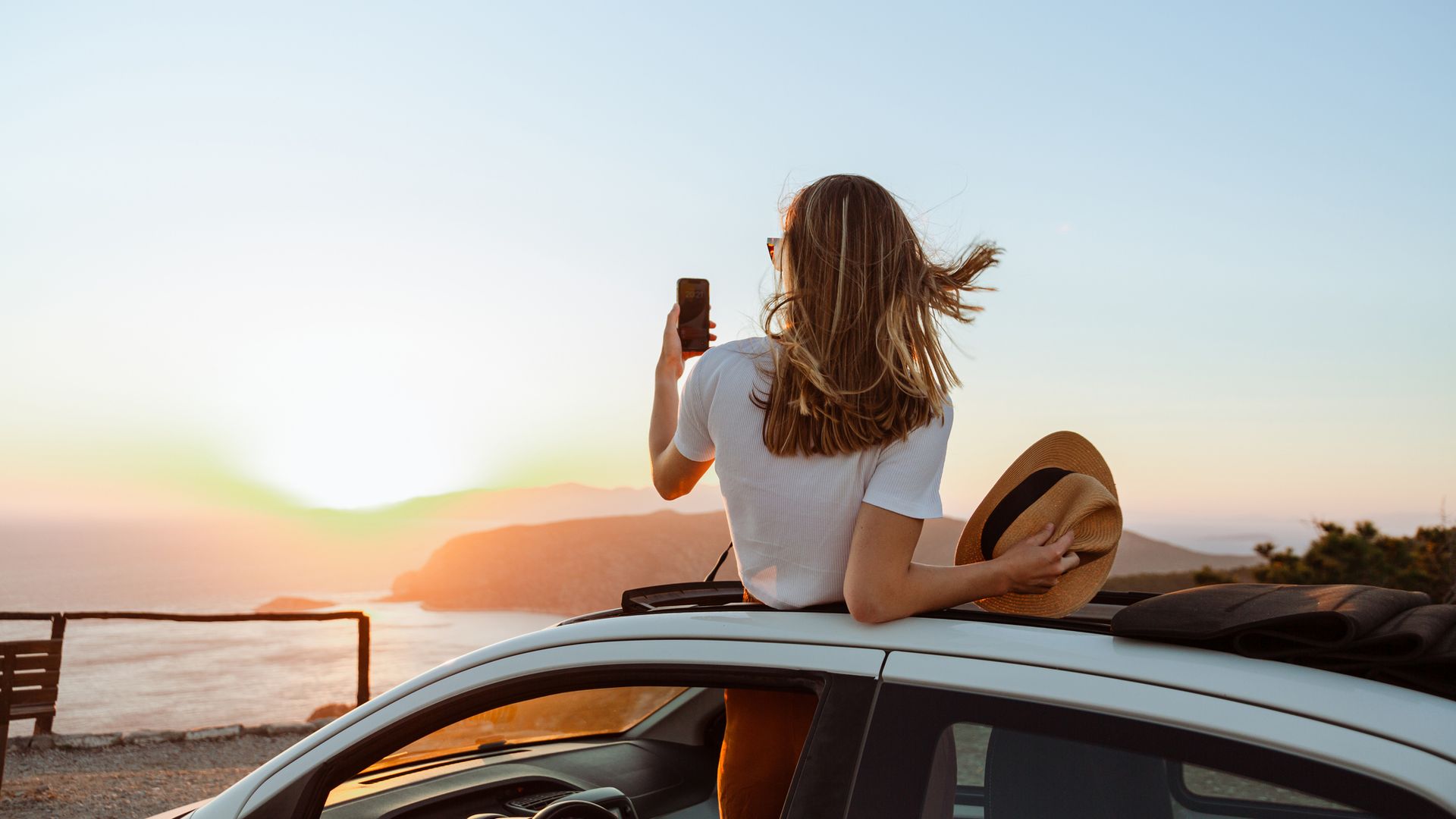 A photo of an unrecognizable woman looking at the sea and mountains from the opened roof of her car. She is taking photos of a beautiful sunset.