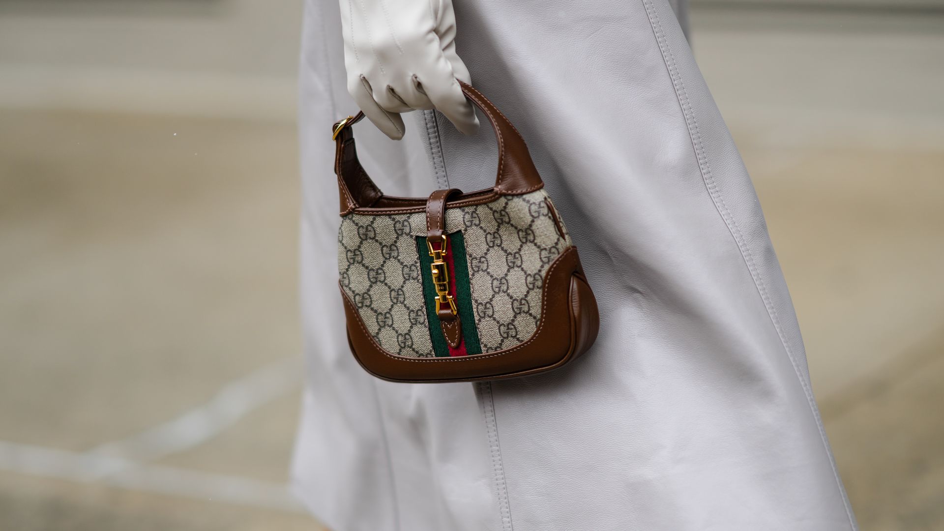 Mary Leest wears a pale gray shiny leather long belted trench coat, white leather gloves, a brown leather with beige and gray GG monogram print pattern handbag from Gucci, orange braided wool high socks, outside Khaite,