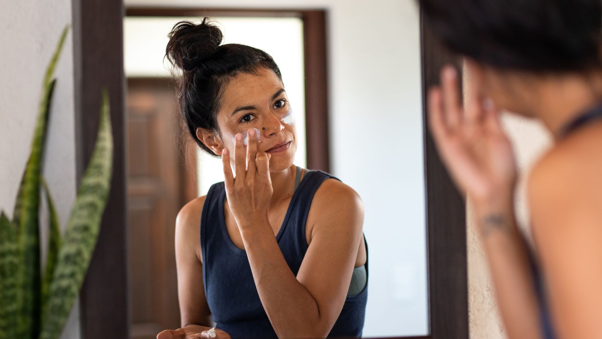 Woman applying white sunblock to face, looking at  bathroom mirror