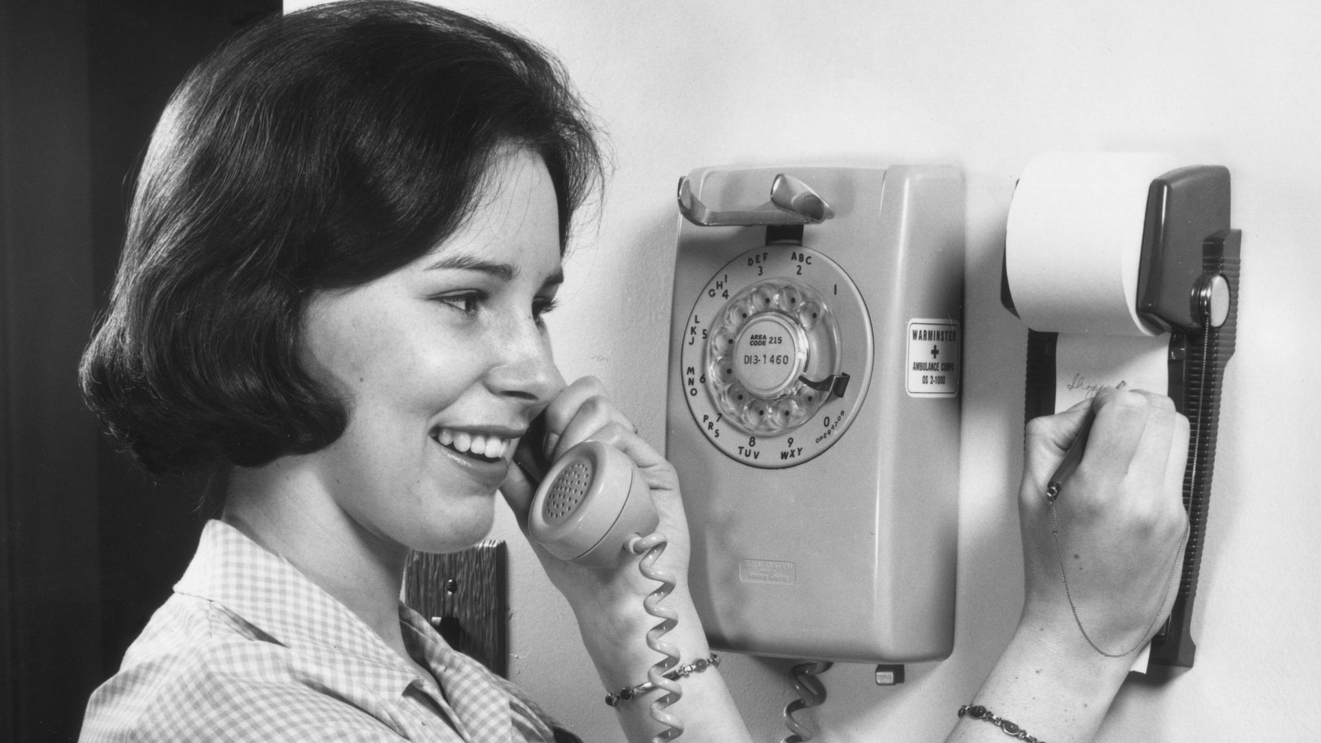 circa 1965:  A woman in a gingham dress talks on a rotary telephone and smiles, as she takes notes on a pad of paper affixed to the wall.  (Photo by Lambert/Getty Images)