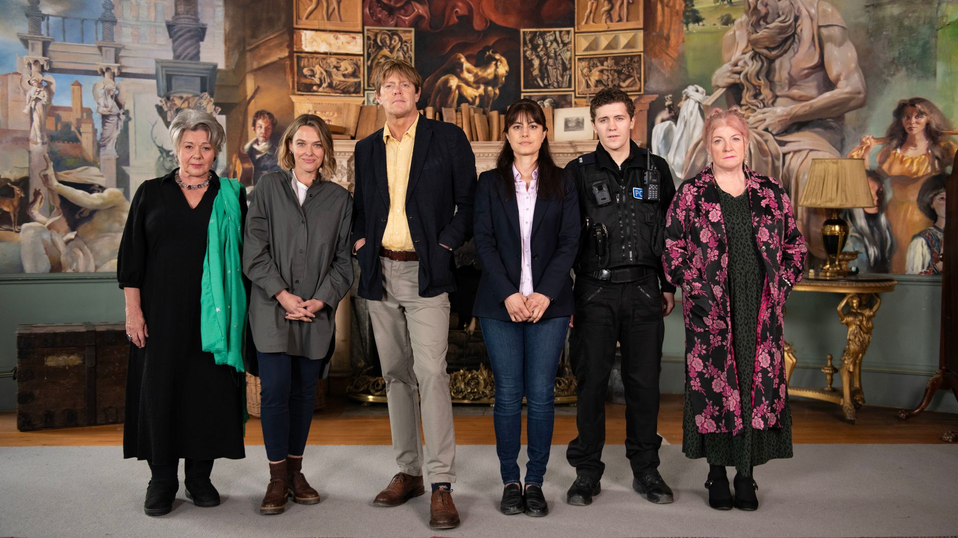 Beyond Paradise cast stand in front of a wall of paintings from episode three. 