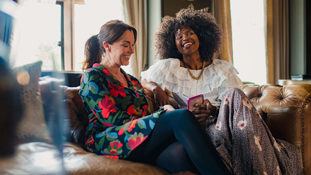 Two beautiful women are sitting comfortably in a luxury hotel, laughing. One of the women is holding a mobile phone in her hand.