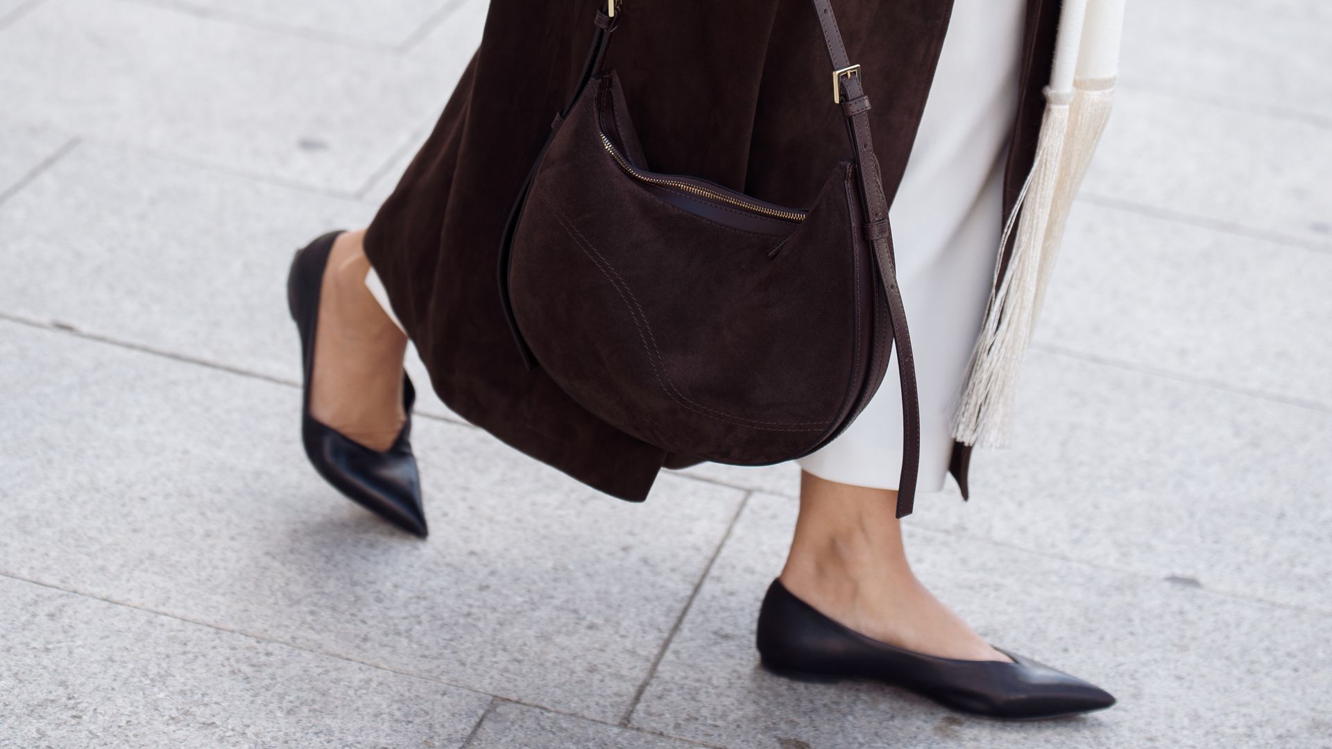 Gili Biegun wears a long chocolate brown suede trench coat, chocolate brown suede shoulder round bag by ATP, cream tassels Scarf, cream midi skirt, flat pointed black leather ballerina shoes outside Matieres Fecales show during the Womenswear Spring Summer 2026