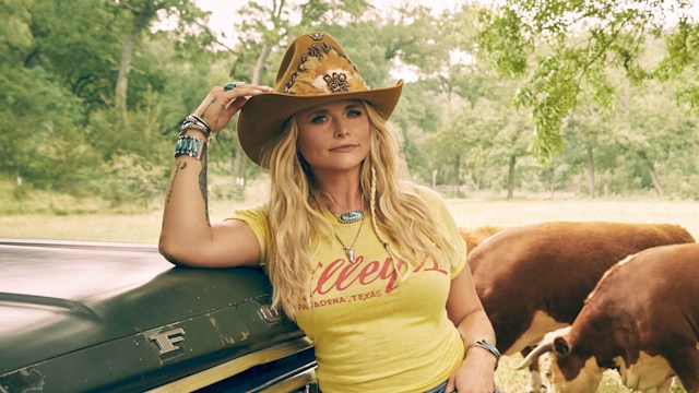 Miranda Lambert poses for picture leaning against truck, four cows stand behind her in a field 