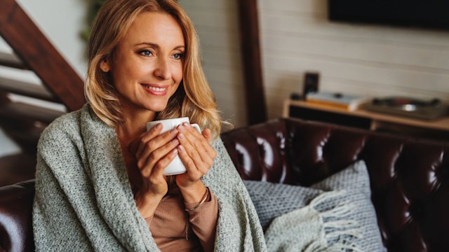 Close up of dreaming middle aged woman sitting in living room with cup of coffee or tea enjoying under blanket