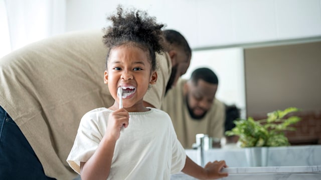 Child brushing her teeth by toothbrush while her father standing behind in the bathroom. 