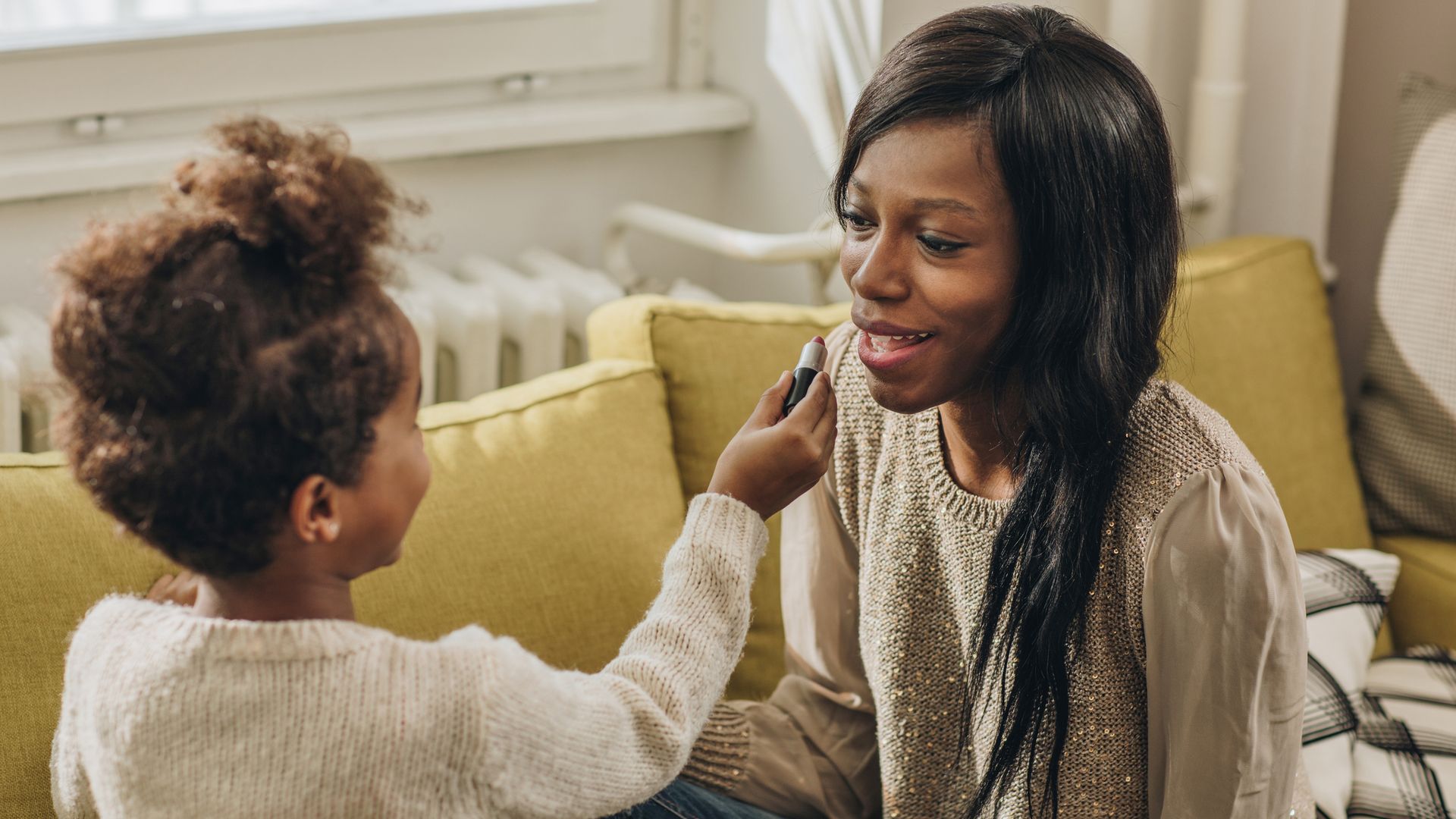 Small black girl having fun with mother at home and applying lip gloss on mother's lips.