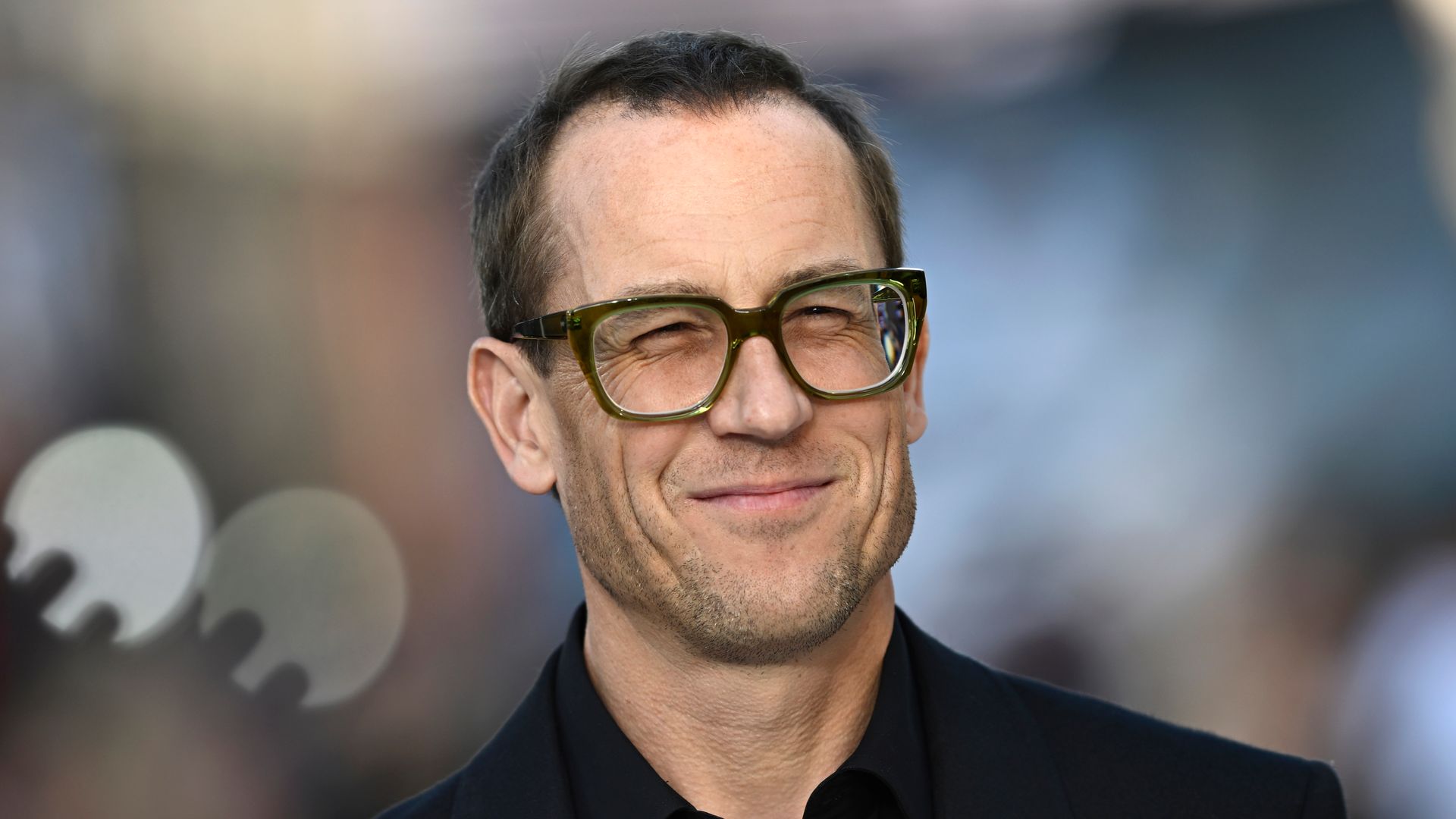 Tobias Menzies squinting in a pair of glasses in a black suit