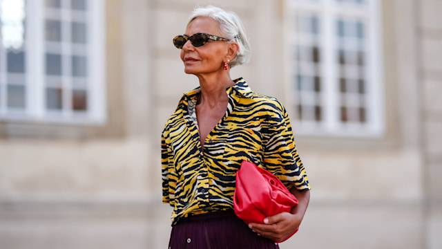 Grece Ghanem wears black sunglasses, red hoop earrings, black yellow and white oversized zebra animal pattern/print short sleeve shirt, shiny red leather bag, outside The Garment, during the Copenhagen Fashion Week Spring/Summer 2024-2025 