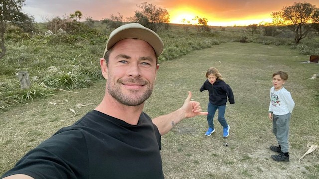 Chris Hemsworth smiling and taking a selfie with his two twin boys behind him playing in a field. The sun is setting behind them.