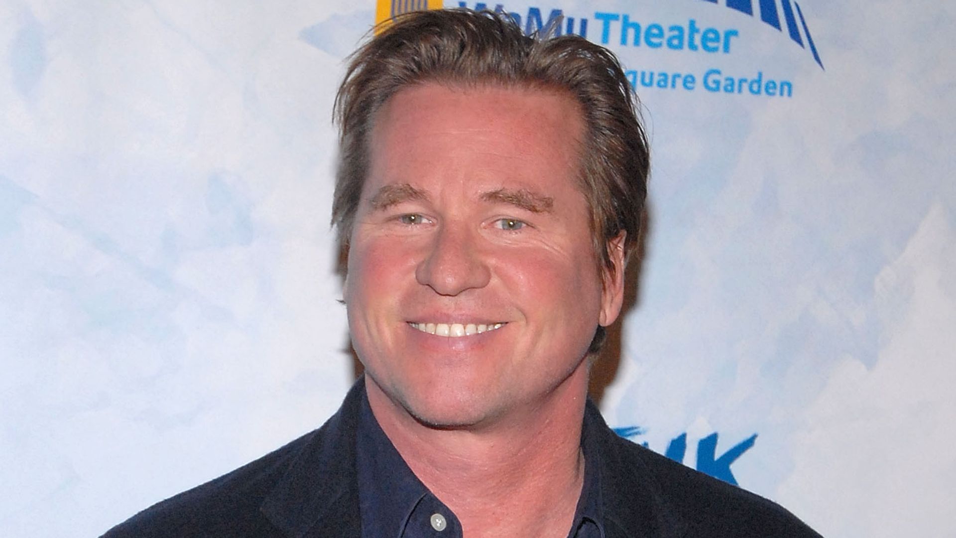 Man standing in front of light blue background in dark blue blazer and navy blue shirt