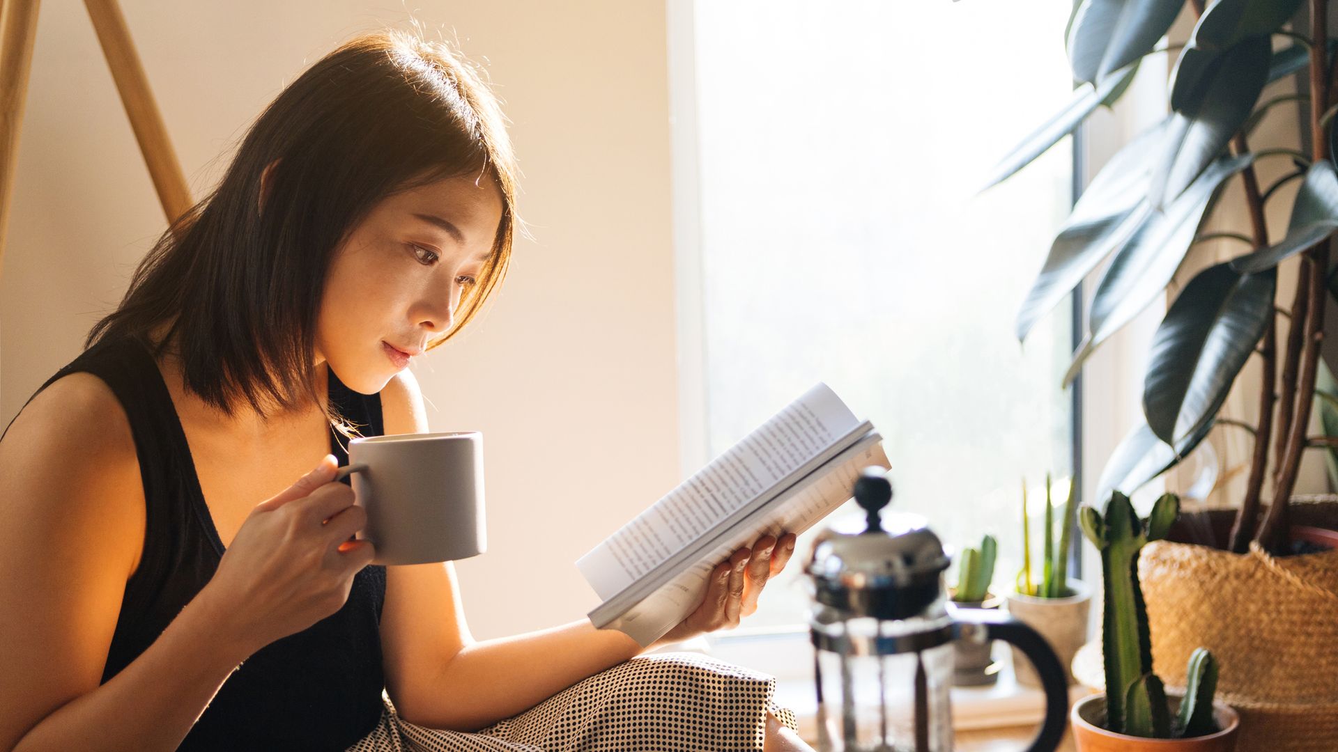 Beautiful young woman reading a book while drinking coffee, enjoying the morning sunlight.