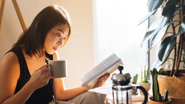 Beautiful young woman reading a book while drinking coffee, enjoying the morning sunlight.