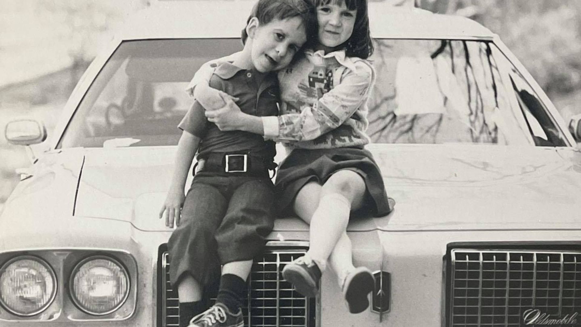 A black and white photo of Ree hugging her brother, they are sat on a car, both are children