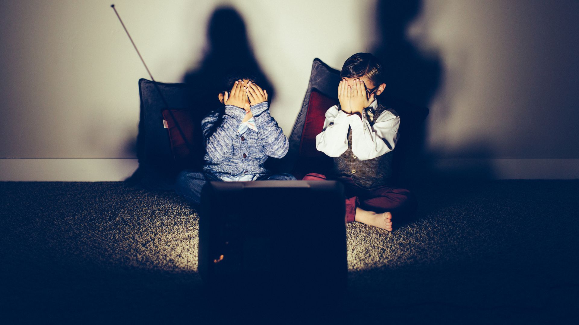 Two young children covering their faces in front of a television