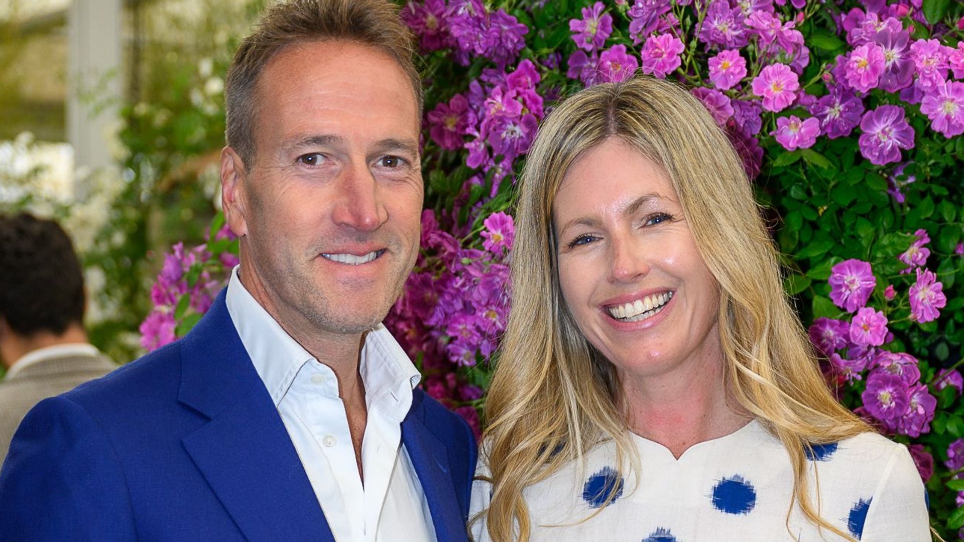 Ben Fogle in a blue suit with wife Marina in front of purple flowers