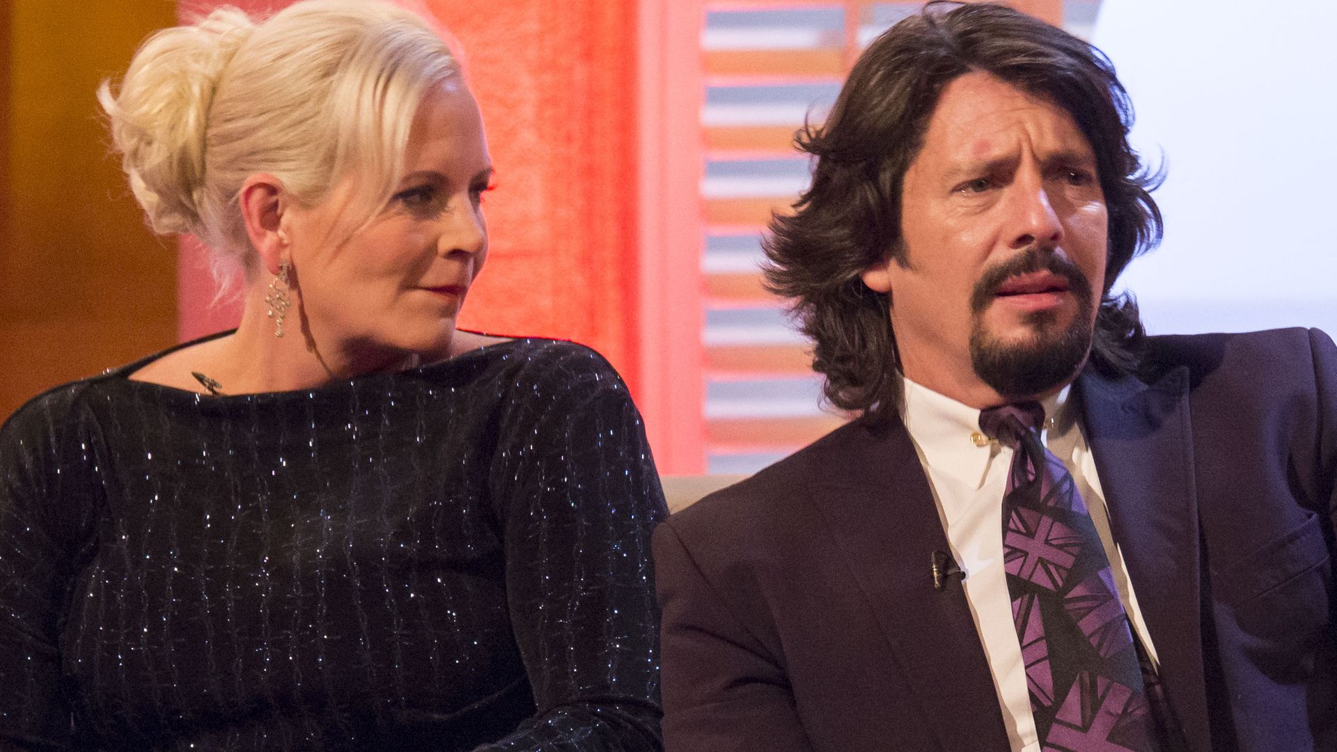 Laurence Llewelyn-Bowen frowning in a suit with his wife Jackie in a sparkly black dress