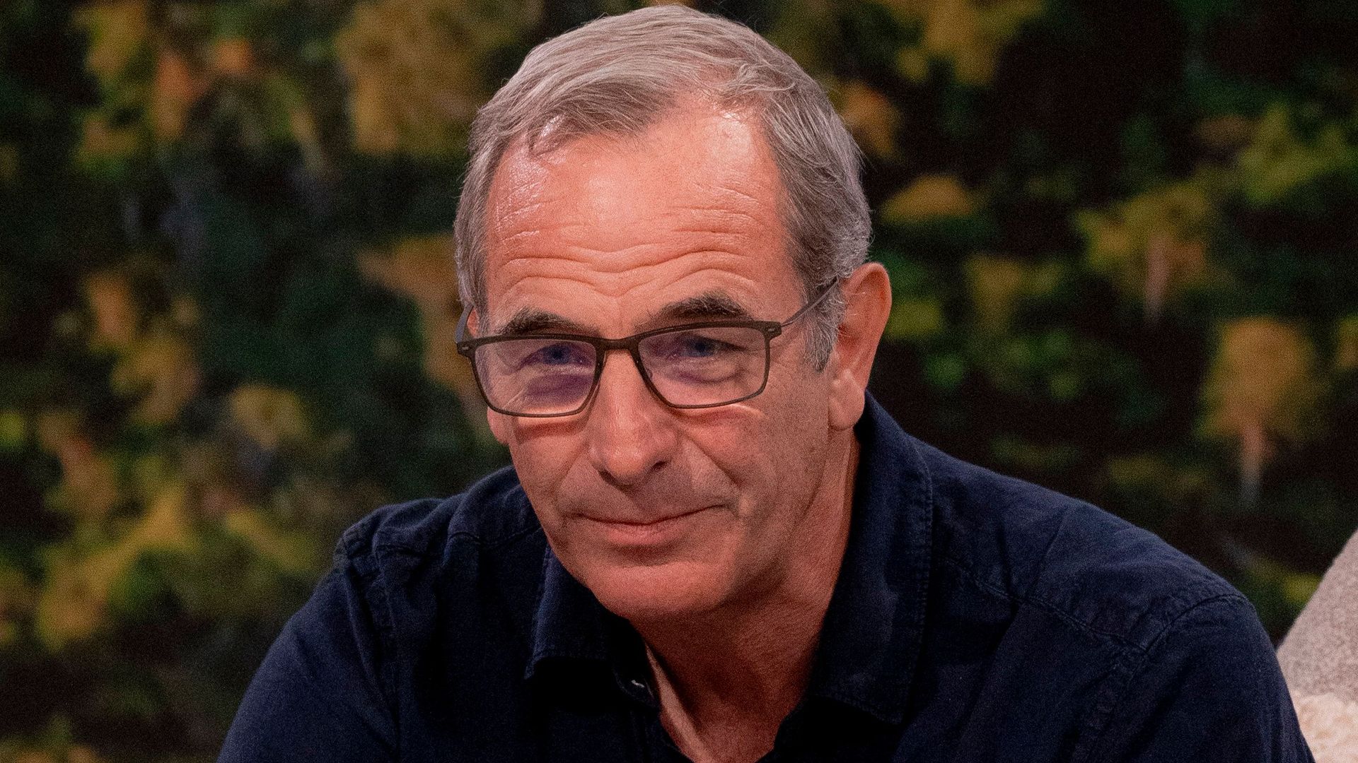 Robson Green in a dark blue shirt in front of a forest 