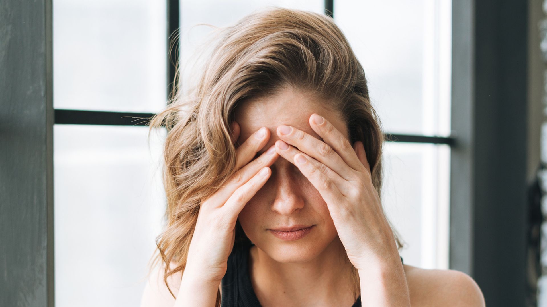 Young brunette woman in black t-shirt covers face with hands near window