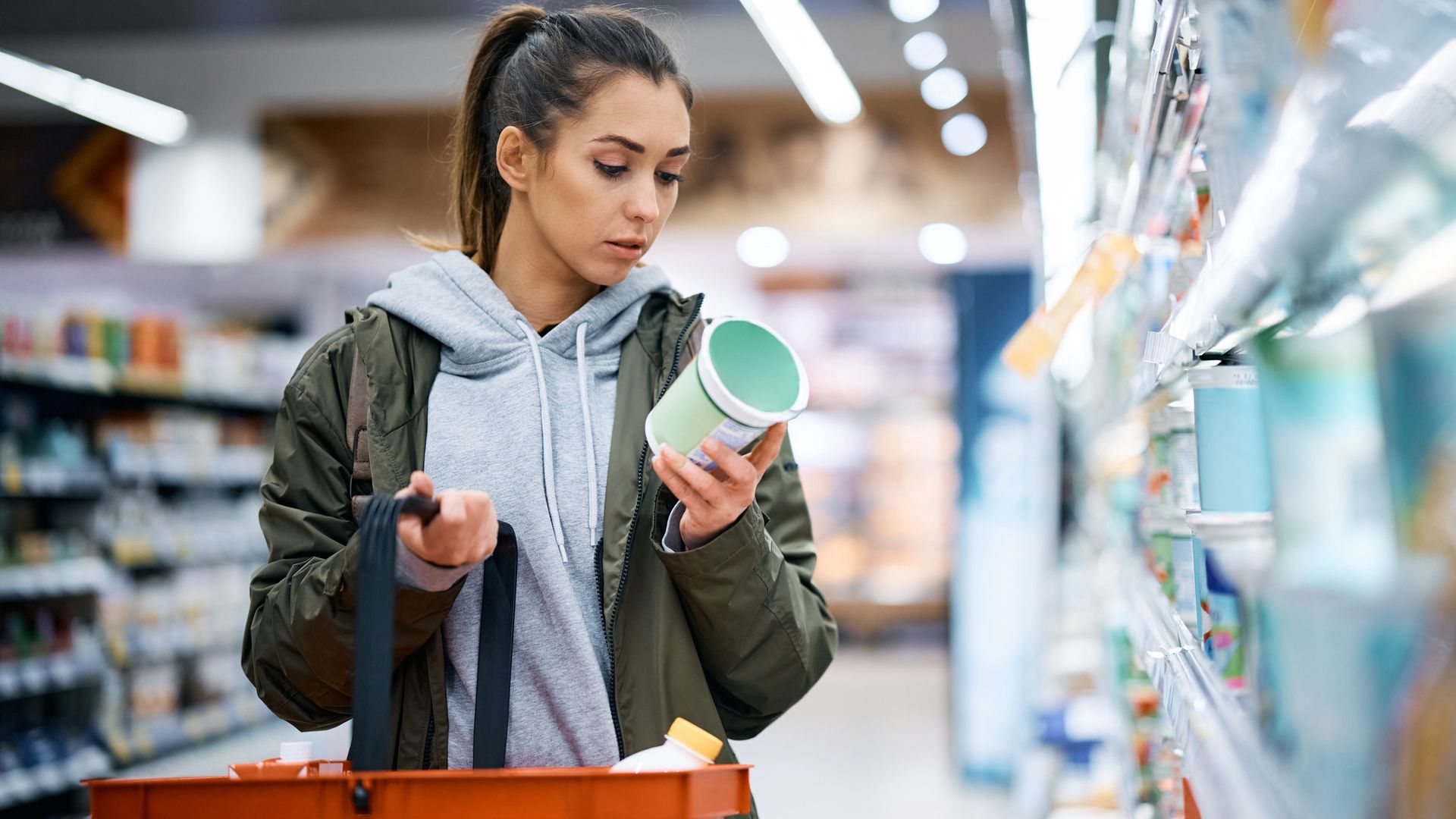 Woman in a grey hoody and green jacket holding a food basket and looking at a dairy food label