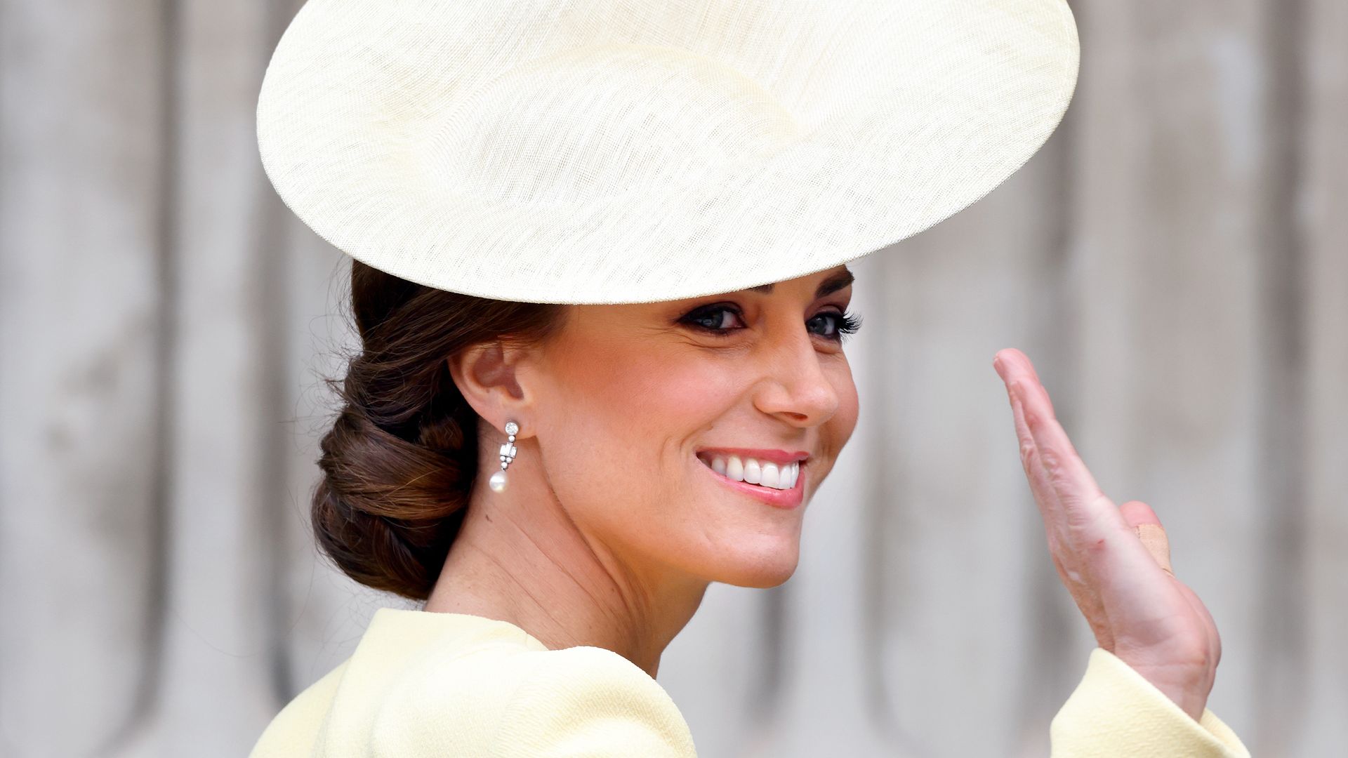Princess Kate waving and looking over her shoulder in a yellow hat and dress