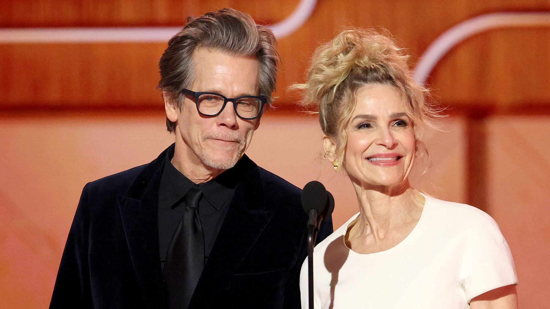 Kevin Bacon and Kyra Sedgwick speak onstage in black suit and white dress