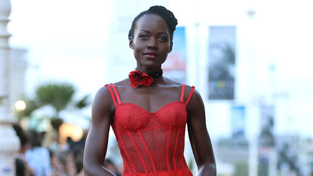 Lupita Nyong'o stands elegantly on the red carpet, wearing a stunning red lace gown with a corset-style bodice and sheer skirt. She holds a small red handbag and accessorizes with a choker and a rose at her neck.