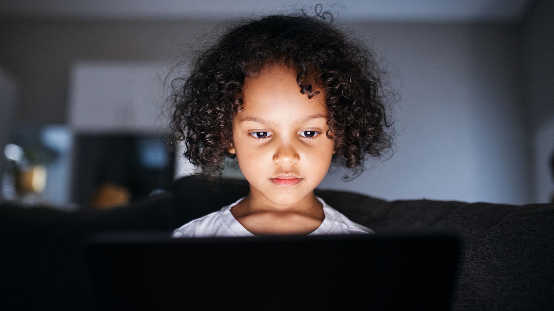 Young girl with curly hair intensely focused on a tablet screen, her face brightly lit by the screen in a dark room. 