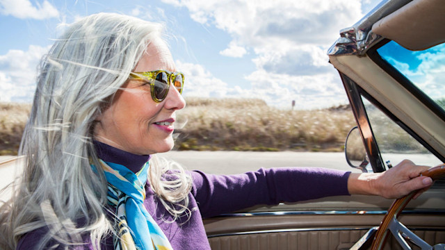  Woman from the baby boomer generation driving a convertible on a sunny fall day with her long, silver gray hair blowing in the wind.