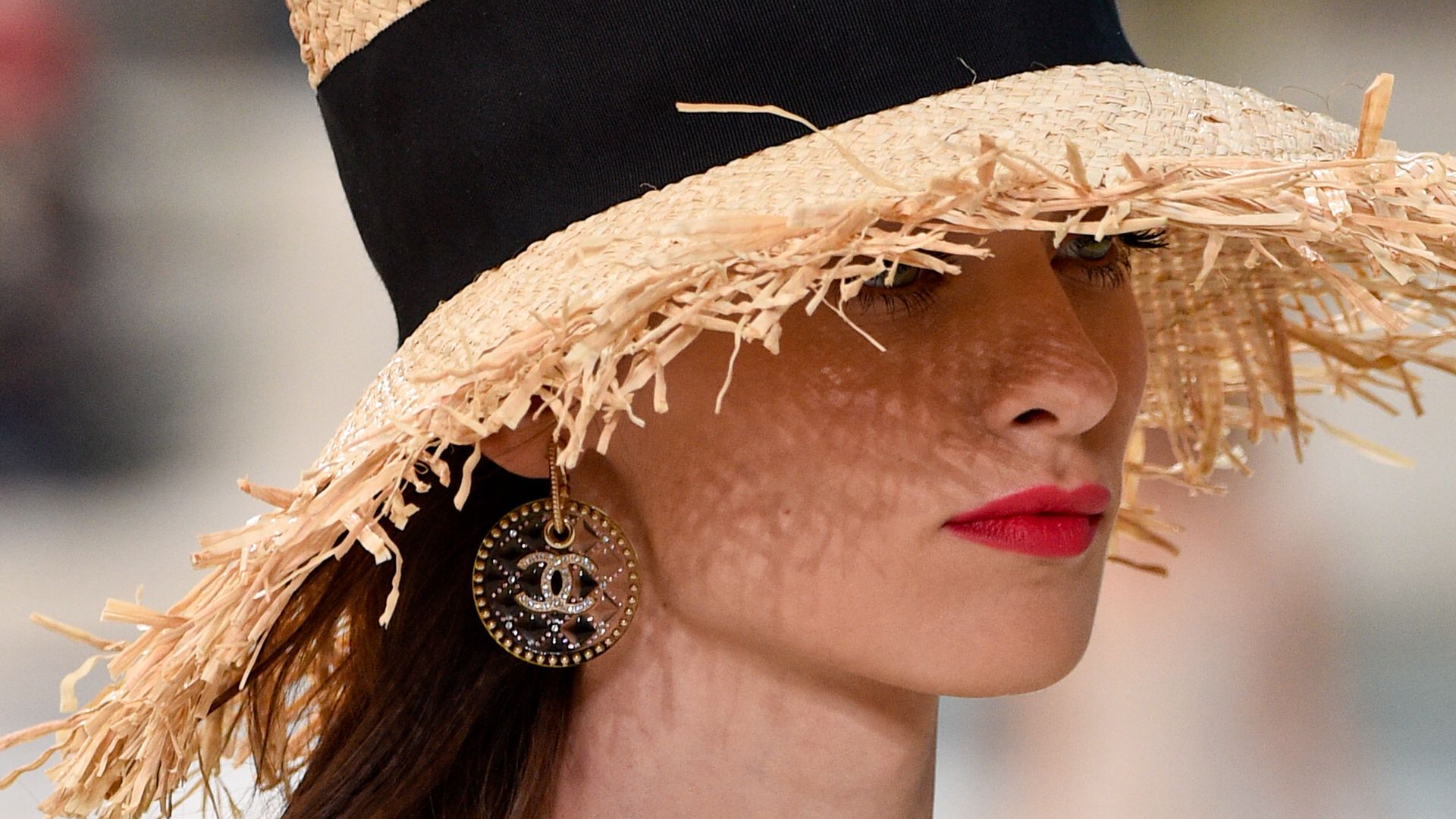 A model wears a wide-brimmed straw hat with frayed edges and a black ribbon, paired with Chanel logo earrings and a gold choker, showcasing a breezy beach look on the Chanel runway.