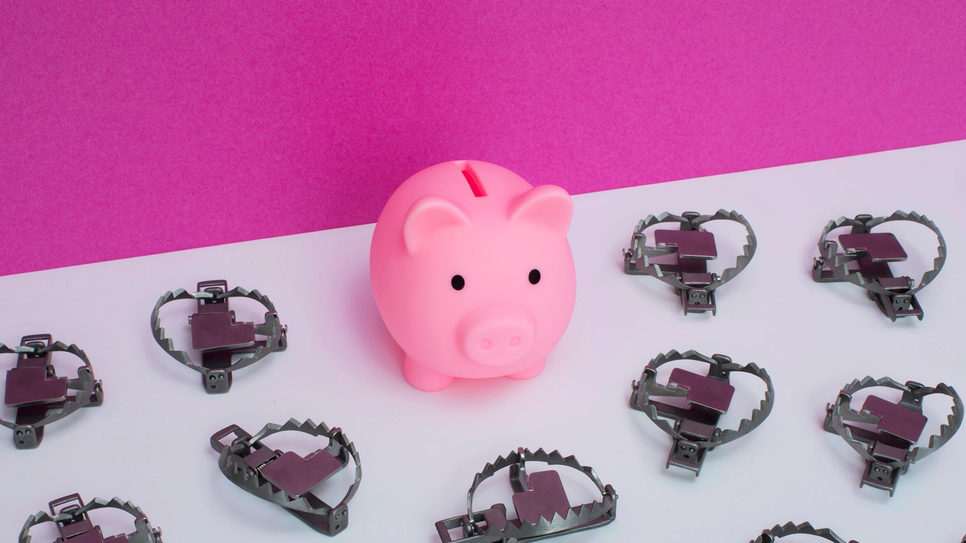 Little pink ceramic piggy bank surrounded by little bear traps on white surface, against pink wall