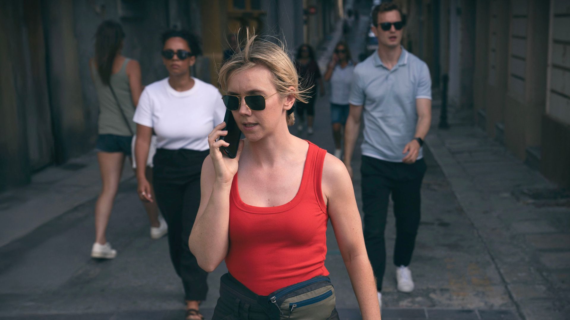 blonde woman on phone in red top walking down street, followed by woman and man in sunglasses