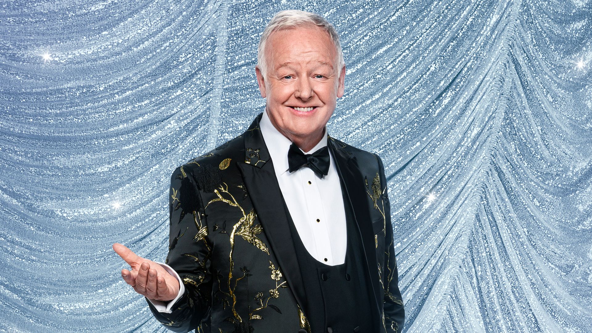 Les Dennis wears a suit and stands in front of blue glittery sheet for Strictly portrait
