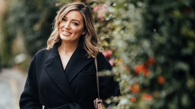 young woman smiling in a black coat standing next to a hedge 