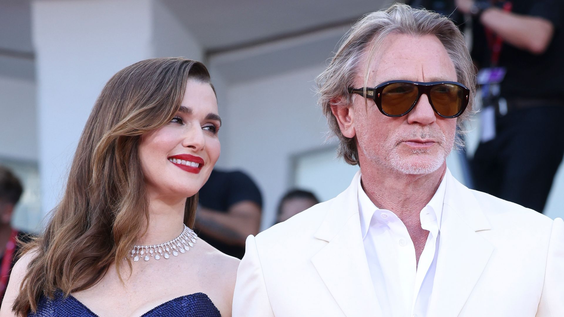 Rachel Weisz in blue dress smiling at Daniel Craig in white suit on red carpet