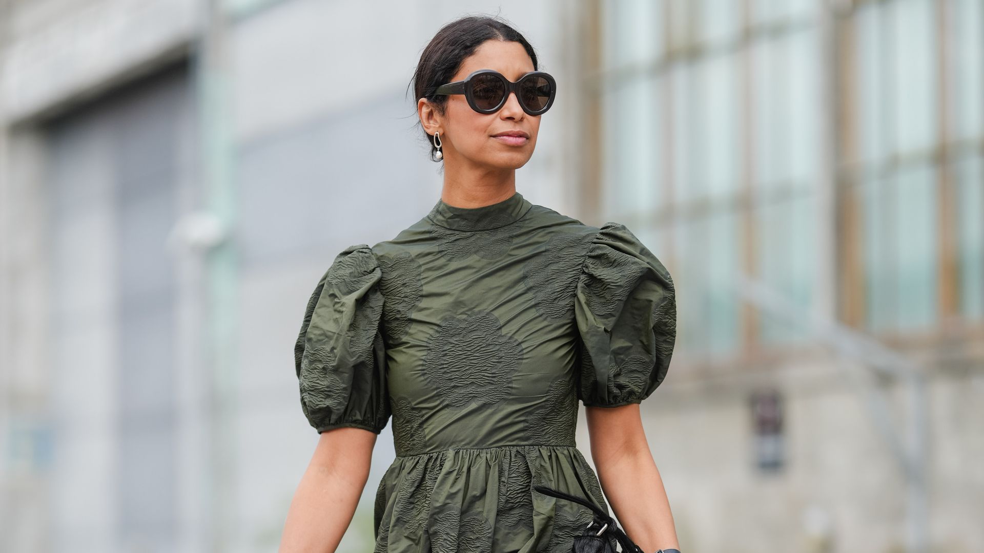 Miriam Nehring wears dark brown hair parted in the center and pulled into a low bun, oversized black rounded cat-eye sunglasses, small goldtone hoop earrings, an olivegreen crinkled midi dress with a high neckline, short voluminous puff sleeves, a gathered waist seam, and a tiered Aline skirt, a blackstrap wristwatch with a round case, a black soft pouch clutch with a ruched top carried in one hand, and black pointedtoe flats with a thin strap and silvertone hardware while walking, outside Marimekko, during Copenhagen Fashion Week
