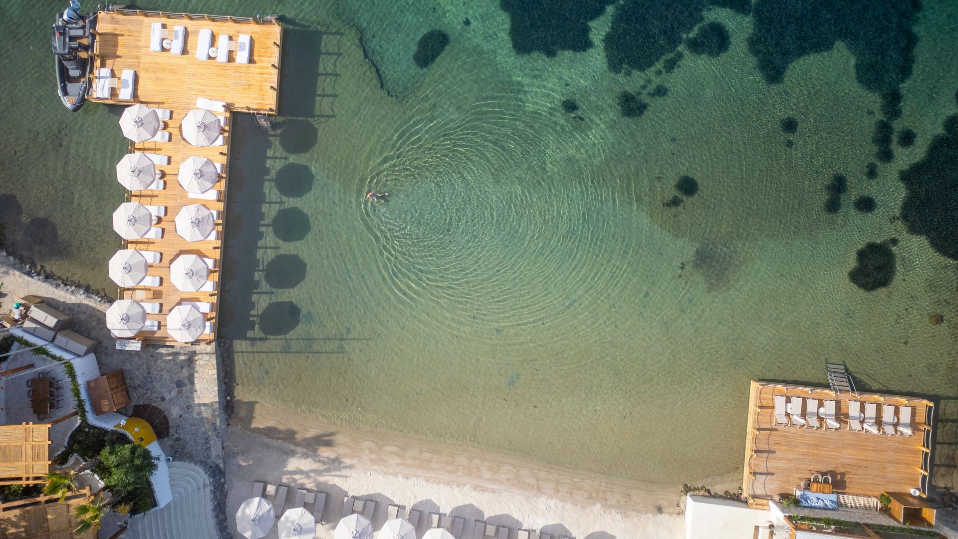 Vue aérienne de la plage privée de l'Avantgarde Refined Bodrum et des terrasses en bois à Yalıkavak, avec des chaises longues blanches, des parasols et des eaux turquoise cristallines le long de la côte égéenne.