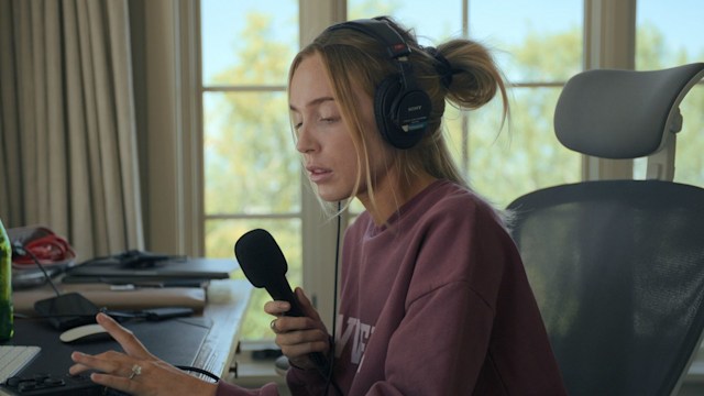 blonde woman holding microphone sitting at desk 