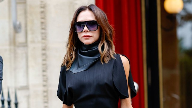 This image shows Victoria Beckham in Paris, wearing a stylish black outfit. She’s in a sleek, short dress paired with black tights and black boots. Her hair is loose, and she’s holding a black handbag, accessorized with large sunglasses and black silk scarf tied around her neck.