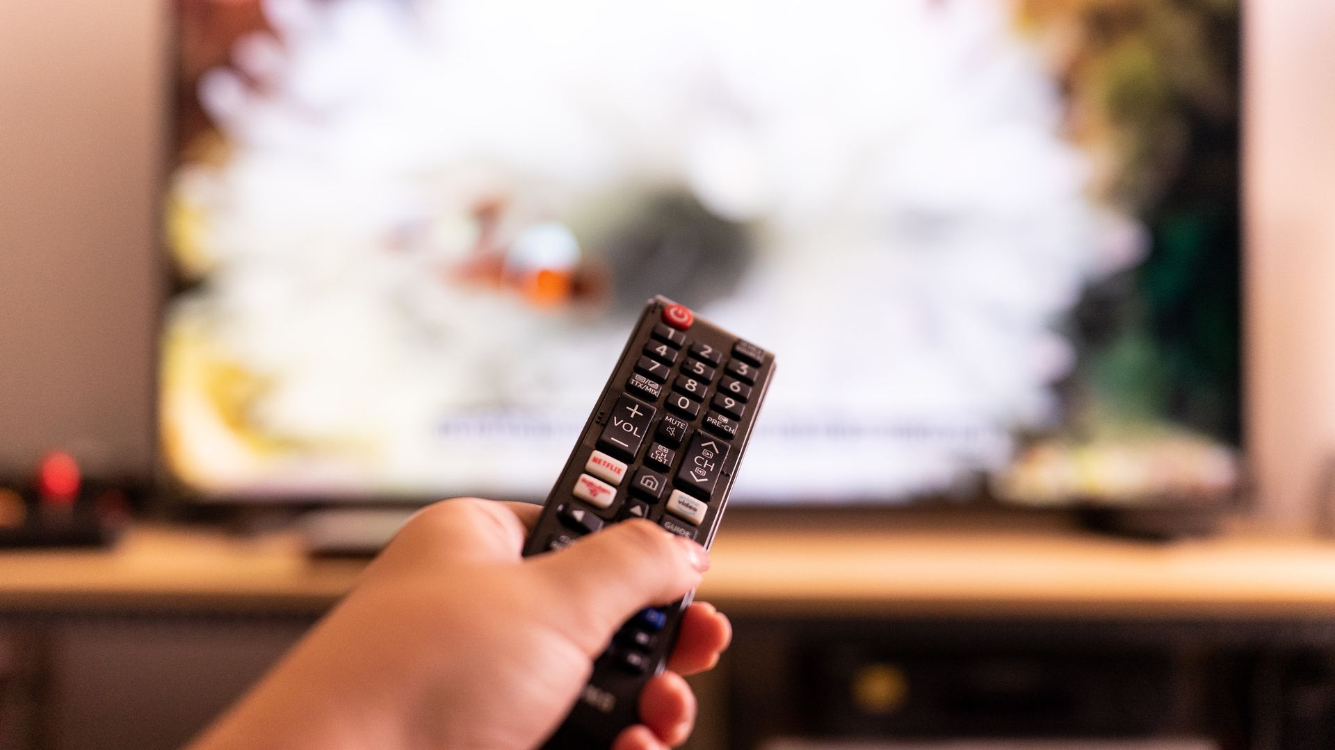 A hand holding a TV remote with shortcut buttons for Netflix, amazon prime video and Rakuten tv streaming platforms, is seen in front of a tv screen